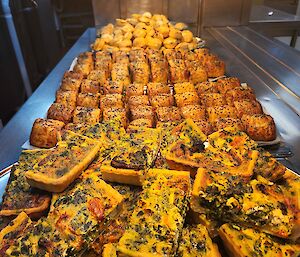 Image is of vegetarian quiche, sausage rolls and party pies stacked up on plates.