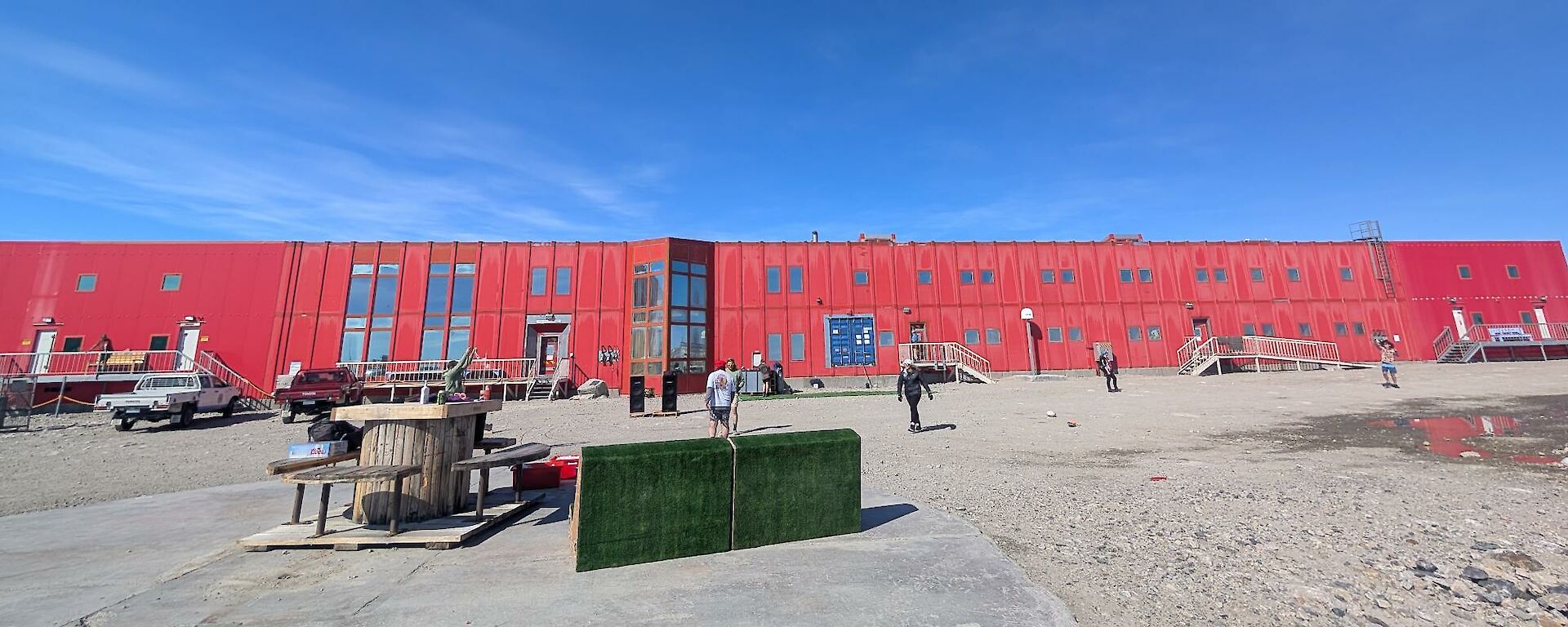 Image shows a group of people playing cricket on a dirt ground infront of the 'Red Shed' with a bright blue sky as a back drop.
