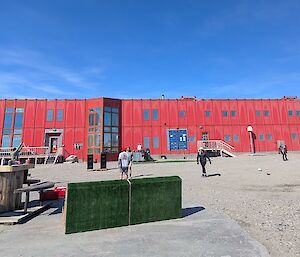 Image shows a group of people playing cricket on a dirt ground infront of the 'Red Shed' with a bright blue sky as a back drop.
