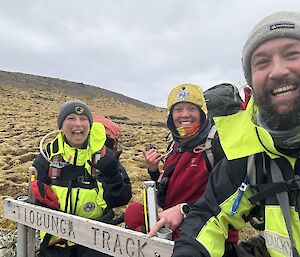 Three smiling people on a hillside with a track sign