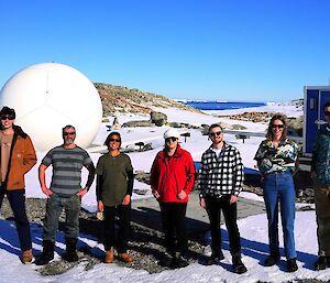Image of 7 people standing abreast. Behind the people meteorological equipment is dotted throughout. The background features the blue sky, ocean and rocks at thge horizon.