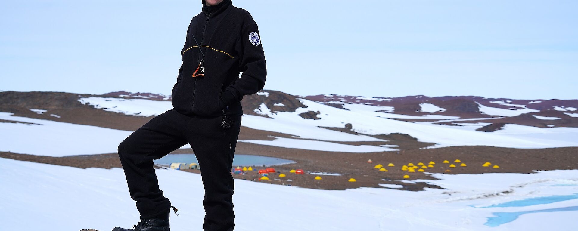 Expeditioner stands on rock in foreground with the Bunger Hills camp in the far background.