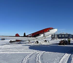 Foreground features snow with vehicle tracks. Mid ground depics a DC3 aircraft (red and white in colour). A blue sky fills the background.