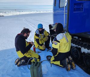 Image of three expeditioners in survival suits attempting to light a stove. Blue Hagglund vehicle is to the right. Snow covered ground below and a blue cloudy sky into the distance.