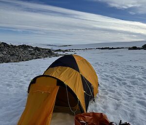 Image of recreation tent set up in snow field. Rock clusters surround with snow covered ground into the distance. Blue sky with cloud in background.