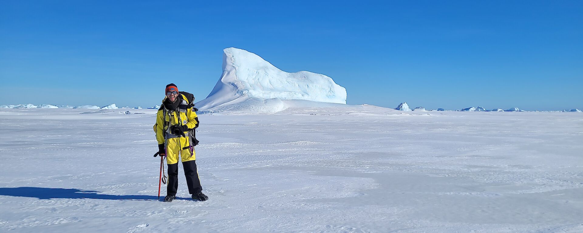 A man standing with an ice axe on ice, with an iceberg behind