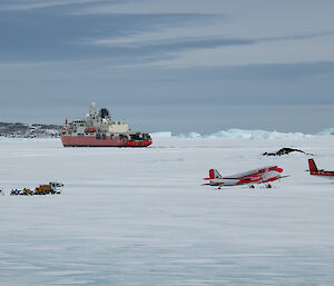 A ship parked in sea ice with two smaller aircraft at front right and some small trucks and quad bikes at front left