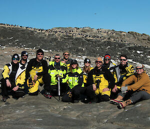A group of expeditioners in high visibility jackets and pants sitting on rocks beneath an Adélie penguin colony
