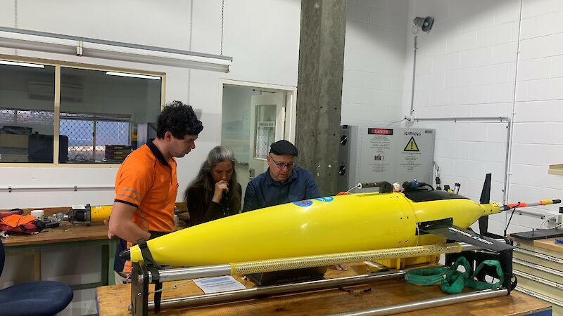 A long yellow metal tube on a table with researchers looking at it
