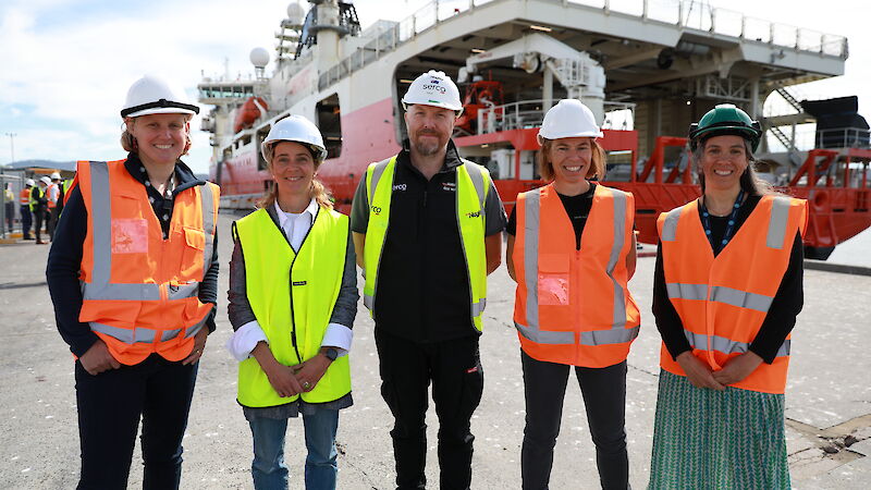 Four men and one man in hi vis on a wharf with a big red and white ship behind them