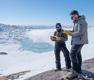 Two men look at hand held tablets, standing on a rocky outcrop in an icy landscape