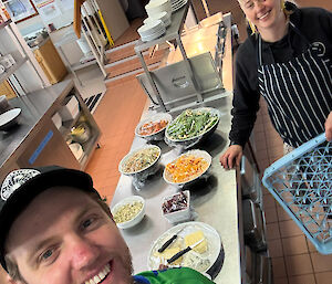 A smiling man takes a selfie with a female chef in a kitchen with food on a table behind him