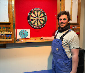 A man in overalls stands in front of a dart board