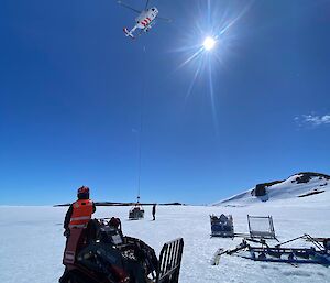 Image of white helicopter in sky with object slung below it. the foreground of image features a person adjacent to a forklift.