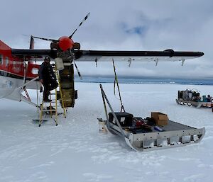 Image of person standing on ladder adjacent to Twin Otter Aircraft with sled positioned nearby.