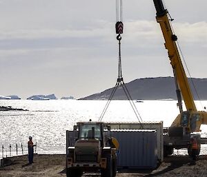 Crane lifts the white metal box with shiny silver bay behind and bare hill