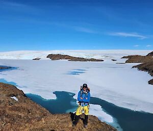 An expeditioner in a blue top and yellow trousers stands on a rocky outcrop with snow going on forever behind him