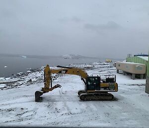 A mustard excavator scoops snow on a flat snowy area with station buildings to the right and a grey sea to the left