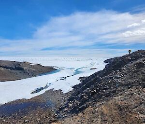 A tiny figure stands on the side of a rocky hill, with a beautiful white and pale green glacier below him