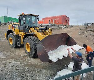 Two people put snow in the scoop of a digger