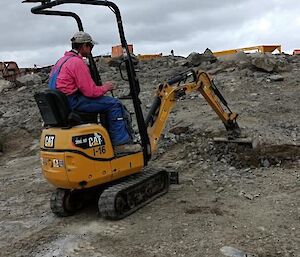 A person in a pink top, cap and blue overalls, drives a very small digger