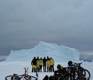 A group of expeditioners stands in front of an iceberg with bicycles in front of them.
