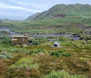 Hut, with power supply and cargo pods, situated in valley with sandy creek behind and large mountains in distance