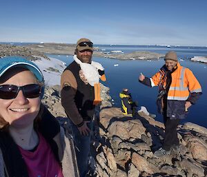 Image of 4 expeditioners with plush toy goose draped around one. Background features ocean and blue sky.