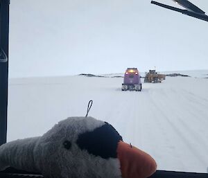 Image of plush toy on dash of Hagg. Background is snow track with Hagg and other plant vehicles driving ahead.