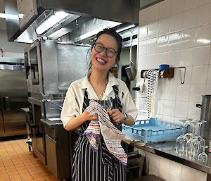 A woman stands smiling in a commercial kitchen, wearing a stripey apron and drying dishes.