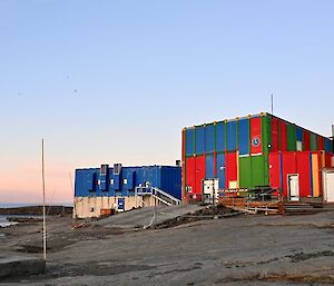 Blue, red and green sheds stand on a rocky slope with a bay behind them