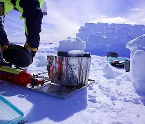Image of an expeditioner using a camping stovein the foreground. The background features a wall of snow built with cut ice bricks.