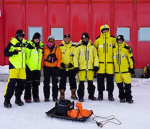 Seven expeditioners standing abreast in their survival uniforms with the red-shed as their backdrop.