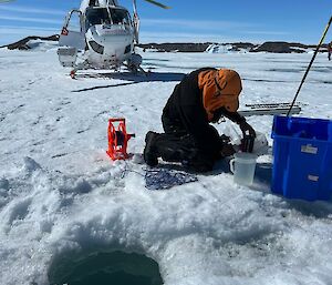Image of a scientist kneeling adjacent to a hole in the sea ice conducting sampling. A helicopter is positioned in the mid-ground with the Bunger Hills rocky landscape in the background.
