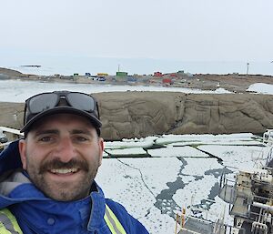 A man looks at the camera with ice and rocks and Mawson station behind him