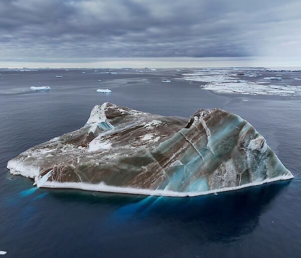A black and green striped iceberg, on flat sea with chunks of ice in the background