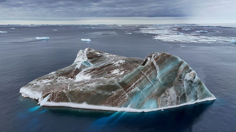 A black and green striped iceberg, on flat sea with chunks of ice in the background