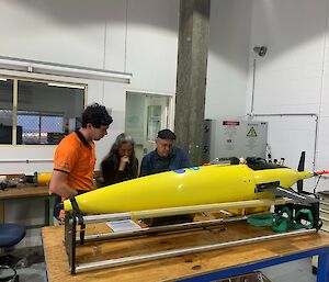 Three people stand looking at a large yellow cylinder on a table