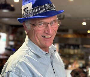 A smiling man in a pale denim shirt and a bright blue hat with sparkles on it.