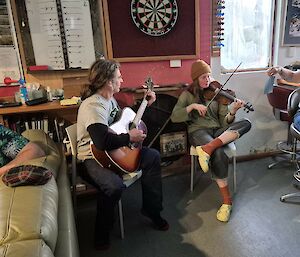 One person on a bar stool with a guitar and a woman sitting on a chair with a violin