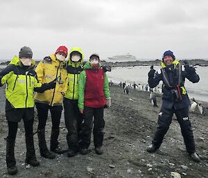 Five people smiling to camera in foreground, behind on beach king penguins and in the distance a ship