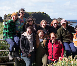 Group of 10 women, 6 sitting on fence, 4 standing in front, smile to camera. In the background a rocky bay.