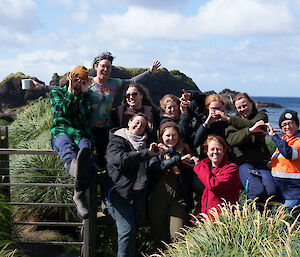 Group of 10 women, 6 sitting on fence, 4 standing in front, make heart signs with hands and smile to camera.
