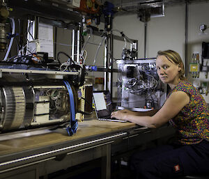 A woman sits at a laptop between two small water-sampling instruments sitting on a bench in a ship's laboratory.