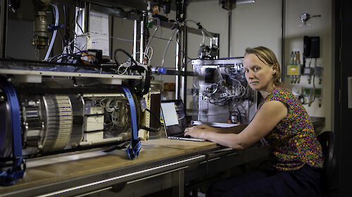 A woman sits at a laptop between two small water-sampling instruments sitting on a bench in a ship's laboratory.