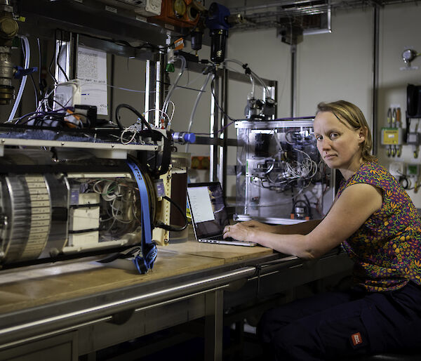 A woman sits at a laptop between two small water-sampling instruments sitting on a bench in a ship's laboratory.