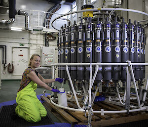 A woman kneels beside a large rosette of bottles. She is holding a small white bottle and collecting seawater from the large rosette using a small length of tubing.
