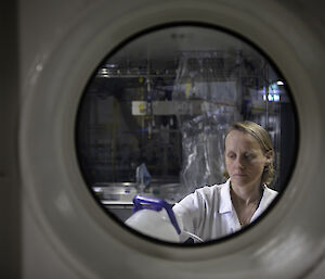 A woman in a white lab coat viewed through a port hole.