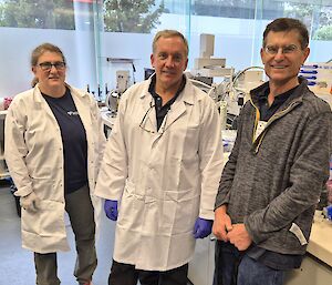 A woman and two men standing beside each other in a laboratory. Two are wearing white lab coats.