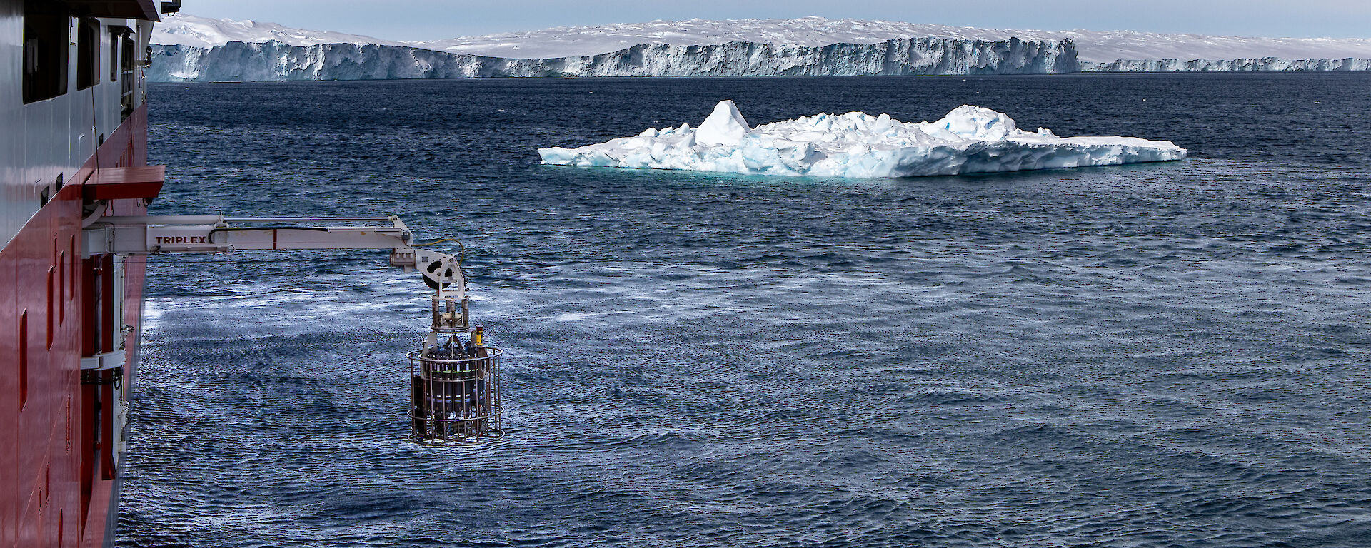 A rosette of water bottles hanging from a small winch from the side of a ship with icebergs in the background.